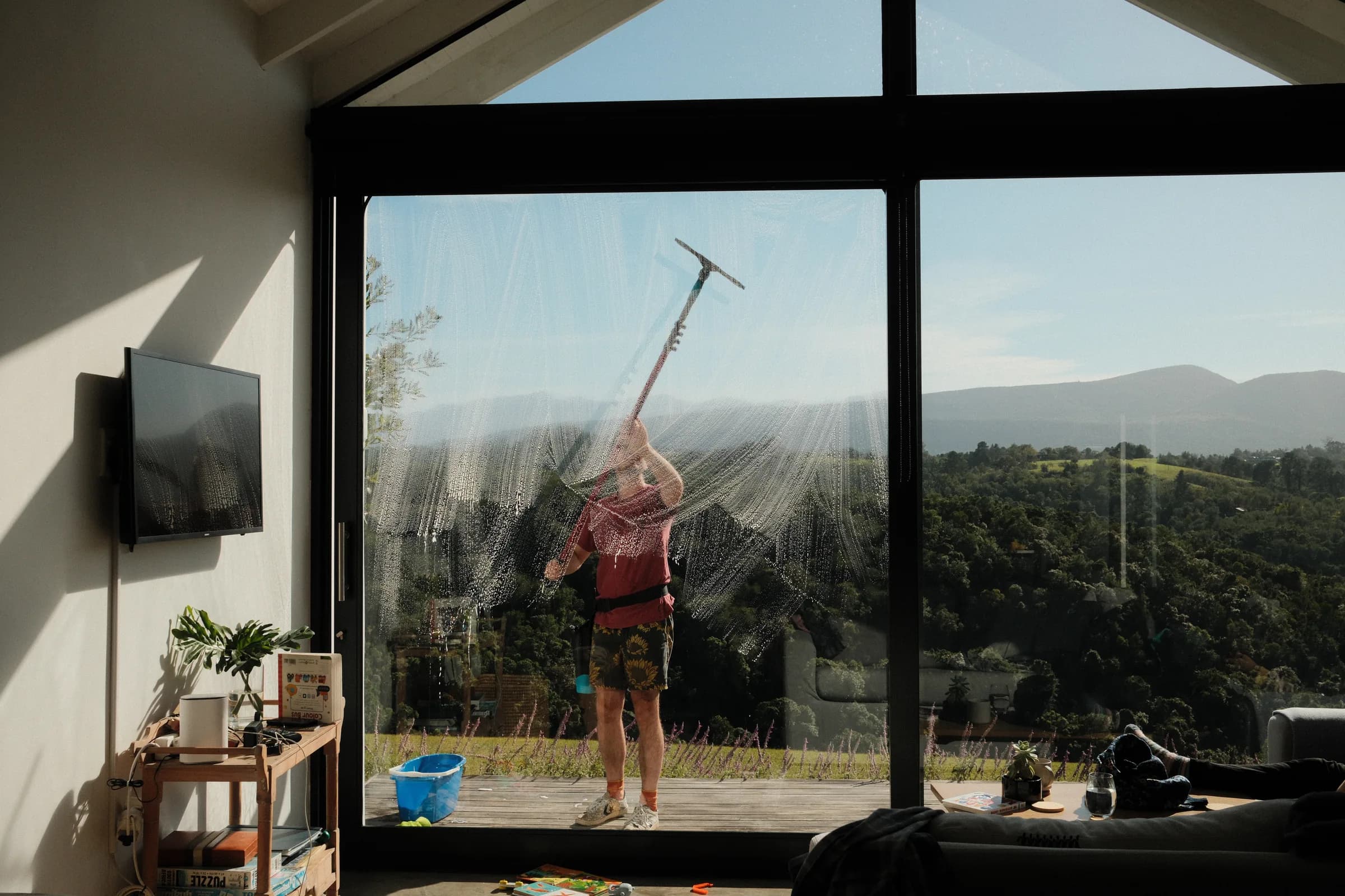 Seen from inside a living room, a ProWash team member squeegees a large glass door with the Knysna hills beyond